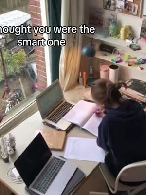 A young woman is seated at a desk cluttered with school supplies and two laptops. She is hunched over a notebook, actively writing. The desk is positioned by a large window overlooking a street scene. The text overlay on the top of the image is in a casual font and reads, 'I thought you were the smart one.' The image has a natural, slightly cluttered aesthetic, suggesting a study environment or workspace.