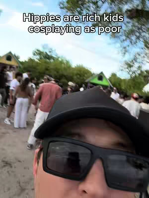 A selfie-style video cover with the upper half of a person wearing sunglasses and a black cap. The person's face takes up most of the frame. In the background, blurry figures of people are seen at an outdoor event or gathering. The sky is visible in the background. A text overlay at the top of the image reads, 'Hippies are rich kids cosplaying as poor'.