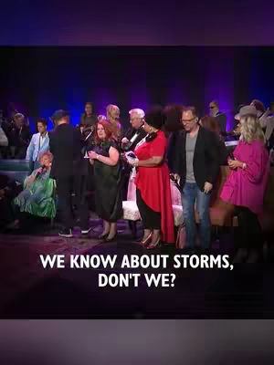 The image shows a stage setting with a group of people standing and some seated in the background. The color scheme is dark with blue and purple lighting. A text overlay in white font near the bottom says, 'WE KNOW ABOUT STORMS, DON'T WE?'