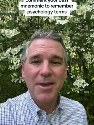 A man with short, graying hair is centered in the frame. He wears a light blue and white checked button-down shirt. Behind him is a backdrop of abundant green foliage with clusters of small white flowers. Above his head, text overlays read, 'Comment your best mnemonic to remember psychology terms'.