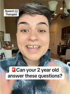 A person with short dark hair smiles widely, revealing a gap in their teeth. They are wearing a dark blue patterned shirt, and the background shows a living room with a chandelier, a side table, and a lamp. Text overlays appear in the image, including the words "Speech Therapist" and "Can your 2 year old answer these questions?".