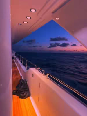 The image captures a serene view from inside a yacht. The foreground shows the side of the yacht with its railing and a glimpse of the wooden deck. In the center, the sky can be seen with its blue color and some clouds at dusk. The water stretches out to the horizon. The lighting suggests either sunrise or sunset.