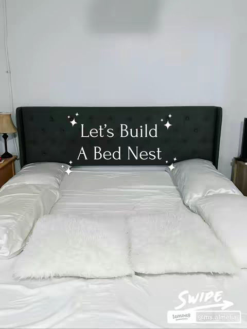 An image of a neatly made bed. The bed has a dark, button-tufted headboard and is covered with white sheets. White pillows are arranged along the sides and at the head of the bed. Two fluffy white pillows sit in the center. The overall impression is of a cozy, inviting bedroom setting.
