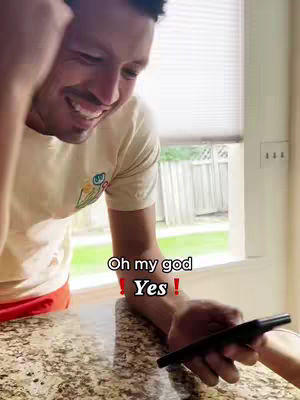 A man smiling widely, holding a phone, with text overlays. He is dressed in a cream shirt, with a view of blinds and and a window to the right of the shot. The photo has a warm, inviting feel.