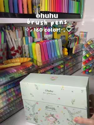 A well-organized display of Ohuhu brush pens is shown on shelves and in clear storage containers. The color palette is extensive with vibrant shades. The text overlay says "ohuhu brush pens 160 color!" with a heart. A box bearing the Ohuhu brand name is also visible.