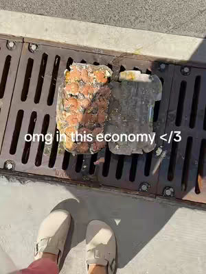 The cover image presents a first-person perspective of a person's feet clad in white shoes, resting on a dark metal grate. Beneath the grate, two containers, one filled with what appears to be eggs, are visible, and the text overlayed says "omg in this economy <3". The scene is lit with natural light casting shadows, and the overall impression is casual and relatable.