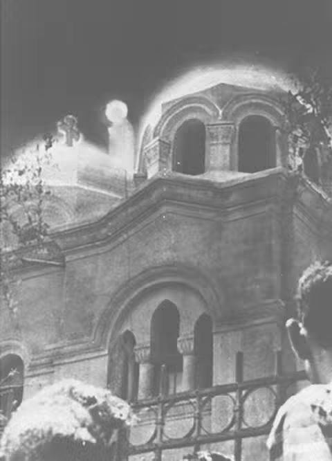 A black and white photo of a church with a domed roof, with an apparition in the sky. People are at the bottom right and bottom left of the frame watching the event. The image has a grainy texture, giving it a vintage feel.