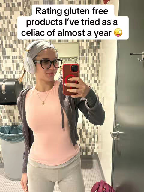 A woman with glasses and a headset takes a selfie in a public restroom. Above her head is a white text box with black text that reads, "Rating gluten free products I've tried as a celiac of almost a year." She has her phone up, which is in a red case, and is standing in a bathroom with a tiled wall.