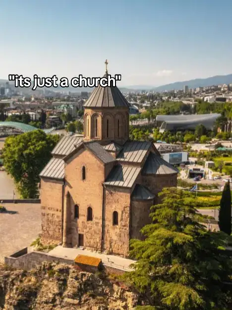 An aerial shot of a church with a brown stone exterior under a clear blue sky. The church has a central dome with a cross, flanked by multiple smaller roof sections. Trees and a cityscape are visible in the background.