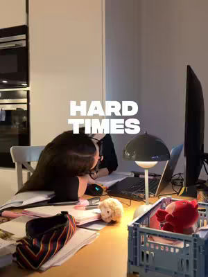 An interior shot shows a woman hunched over a desk, presumably studying or working, with a simple, modern aesthetic. Visible elements include a laptop, lamp, papers, and some small decorative items. The lighting is warm, setting a cozy, studious atmosphere. The composition is centered, with the focus on the woman and her workspace.