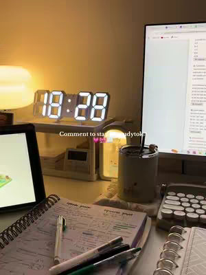 A well-lit desk scene with a digital clock prominently displayed. A laptop is on the left, and notes and pens are in the foreground. Soft lighting gives a cozy, studious vibe