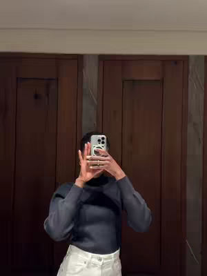 A person, appearing to be a woman, is taking a selfie in a mirror. The person is wearing a dark top and white pants, with arms raised to hold a phone in front of the face. The background includes wooden doors and a neutral-colored wall. The photo has a vertical composition.