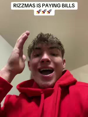 A young man with light brown hair and a red hoodie is shown in a selfie-style video. He is smiling and gesturing with his right hand raised, while his mouth is open as if he is speaking or laughing. In the top, there is a text overlay with the phrase "RIZZMAS IS PAYING BILLS" and three rocket emojis.