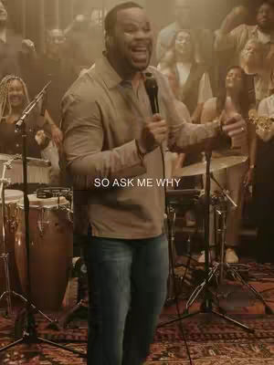 A man wearing a light brown button-down shirt and blue jeans is in the foreground, holding a microphone and singing, with a large choir behind him and musical equipment on the sides. The background is softly lit to add depth.
