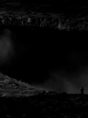 A grayscale image featuring a dark, imposing landscape. The top portion is dominated by a rock formation, possibly a cliff face or a large rock. Below, a dark, misty area suggests water or fog. Near the bottom right, a small, silhouetted figure of a person stands, offering a sense of scale and perspective against the vastness of the scene. The overall composition is moody and stark, with high contrast between light and shadow.