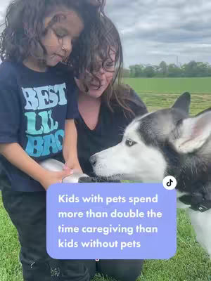 A young child and a woman are embracing a dog outdoors. The child is wearing a dark blue t-shirt with white and teal block lettering. The woman wears a black v-neck. A large, black and white husky is positioned on the right side of the frame. A light blue rectangular text overlay displays a message.