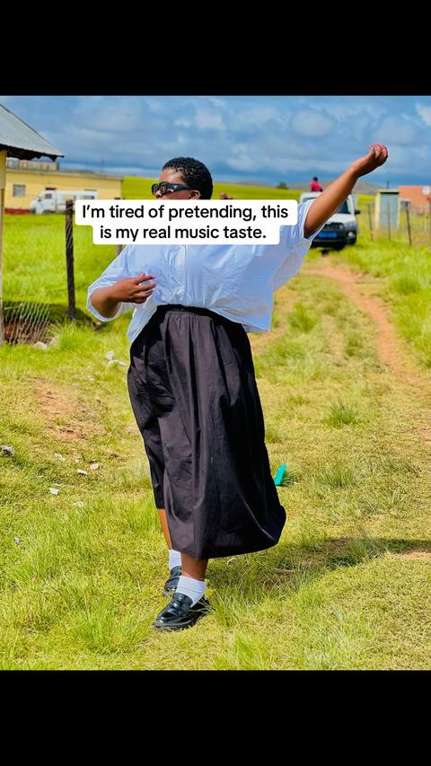 An individual, likely a woman, wearing sunglasses and is gesturing with one arm raised towards the sky. She is dressed in a white, loose-fitting shirt and a long, black skirt, standing in a grassy, outdoor area with a path and a building in the background.