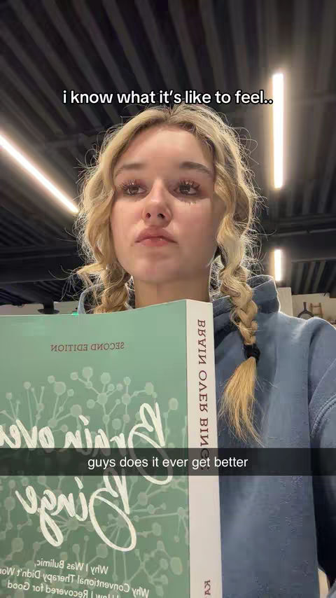 A young woman with blonde pigtails holds a book in front of her, looking toward the camera with a somber expression. She is inside a building with a gray ceiling and vertical lights. A text overlay says, 'i know what it's like to feel' and 'guys does it ever get better'.