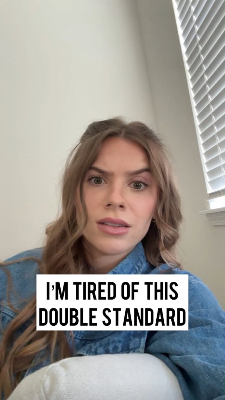 A woman with long wavy brown hair is looking at the camera with a slightly concerned expression. She is wearing a denim jacket. A white text box sits in the lower center, with the text "I'M TIRED OF THIS DOUBLE STANDARD". The background is a white wall, with a window and blinds visible to the right.