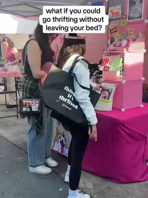 The image shows three women standing at a pink table in what appears to be a thrift store. Text overlay reads 'what if you could go thrifting without leaving your bed?'.