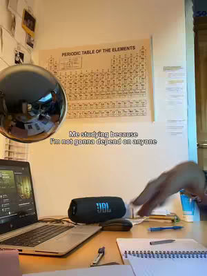 The image shows a room set up as a study or workspace. A laptop and a JBL speaker sit on a desk. Behind them is a periodic table of elements chart hanging on the wall. The person in the study appears to be reaching for something on the desk, visible in the frame. The overall composition suggests a study or work environment.