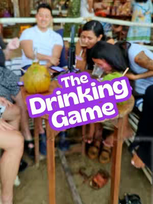 A group of people are gathered around a table with drinks. A large purple banner labeled 'The Drinking Game' is superimposed on the image. The background is blurred, suggesting an outdoor or casual setting.