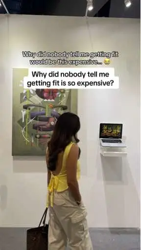 A woman stands in a gallery, turned away from the viewer, looking at a painting and a laptop mounted on the wall. She wears a yellow top and beige pants and has her hair down. Text overlays on the image.
