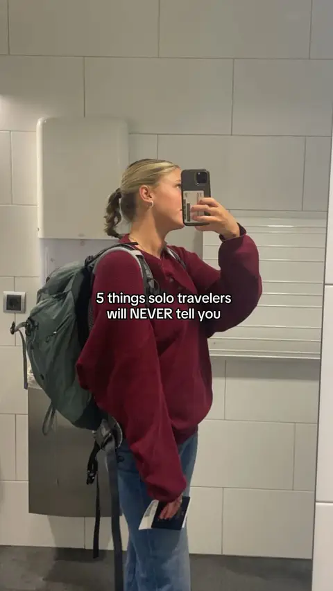 A woman is standing in a bathroom looking at her phone, holding a passport. She's wearing a red sweatshirt with jeans and has a backpack. The background shows white tiled walls and a mirror.