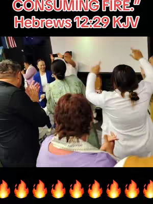 The image shows a group of people in a lively scene likely representing a church service or a gathering. At the top of the image is the hook text, which is in a contrasting red color and reads "CONSUMING FIRE." followed by "Hebrews 12:29 KJV". The scene is a medium shot with a shallow focus, with a group of people visible dancing around the two women who are in the center of the shot. At the bottom is a black bar with orange flame icons.