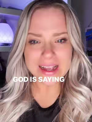 A close-up shot of a woman with long blonde hair, wearing a black top. The woman appears to be emotional with wet eyes and an open mouth. Overlayed on the image is white text reading, "GOD IS SAYING."
