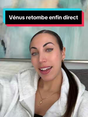 A woman with dark hair in a low ponytail, wearing a white hoodie and a gold necklace, speaks directly to the camera. A text overlay in a colored banner says, "Vénus retombe enfin direct". The background is a blurred interior with a painting.