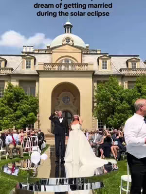 A wedding ceremony is depicted outdoors, with a grand building in the background under a clear, blue sky. In the foreground, a mirrored platform reflects the couple, who are dressed in wedding attire, and the surrounding guests. Trees and seating arrangements add context to the event.
