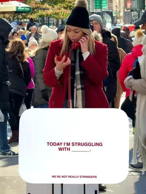 The cover image shows a woman on a crowded street. She is holding a red phone up to her ear. A white banner at the bottom features the text: "TODAY I'M STRUGGLING WITH ______" and "WE'RE NOT REALLY STRANGERS". She is wearing a black beanie with a fur pom-pom, a red coat, a scarf, and is surrounded by people, creating a sense of being in public.