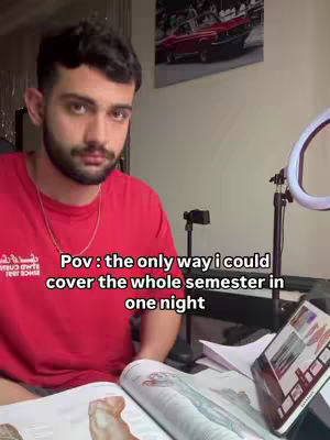 A young man with a beard and dark hair is sitting at what looks like a desk with several items around him and a ring light. He is wearing a red t-shirt and has text overlaid on the bottom of the image.