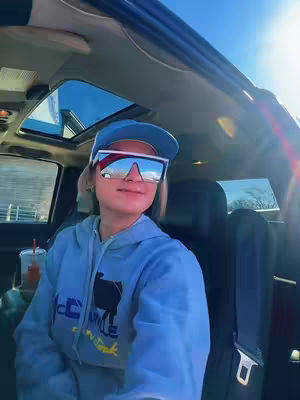 A person wearing large reflective sunglasses and a blue hoodie is seated inside a car. The interior has a sky-blue tinted glass roof and side window, with the sunny exterior reflected in both the sunglasses and the car's tinted glass. The person has short, light-colored hair. A partially visible drink is in the foreground.