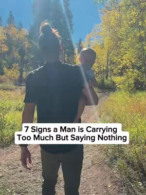 A man is walking on a dirt path in a forest, carrying a child. The man is seen from the back, wearing a dark t-shirt and cargo pants. The child is wearing a light blue shirt. The background is a blur of green trees with yellow and orange leaves, under a clear blue sky.