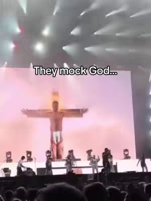 The image shows a stage performance with a figure in the middle, resembling Jesus on a cross. The stage is brightly lit with spotlights and has people on stage in front of a large screen. The text overlay at the top says, 'They mock God...'