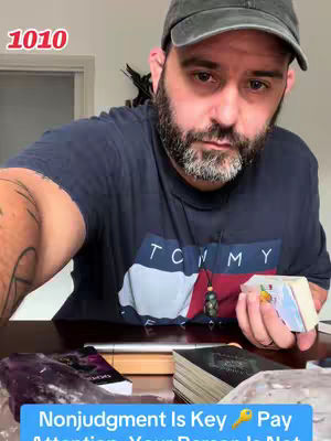 A man with a beard and a baseball cap is looking into the camera. There is text overlaid on the bottom of the image. There are tarot cards on a table in front of him. There is an amethyst in the lower left corner.