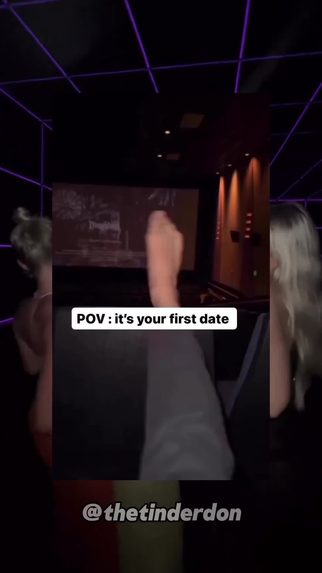 The image shows a POV shot filmed in a movie theater. The scene focuses on a person's crossed legs and another figure. In the background, a movie screen is visible, along with the interior of the theater, including seating arrangements and lighting.