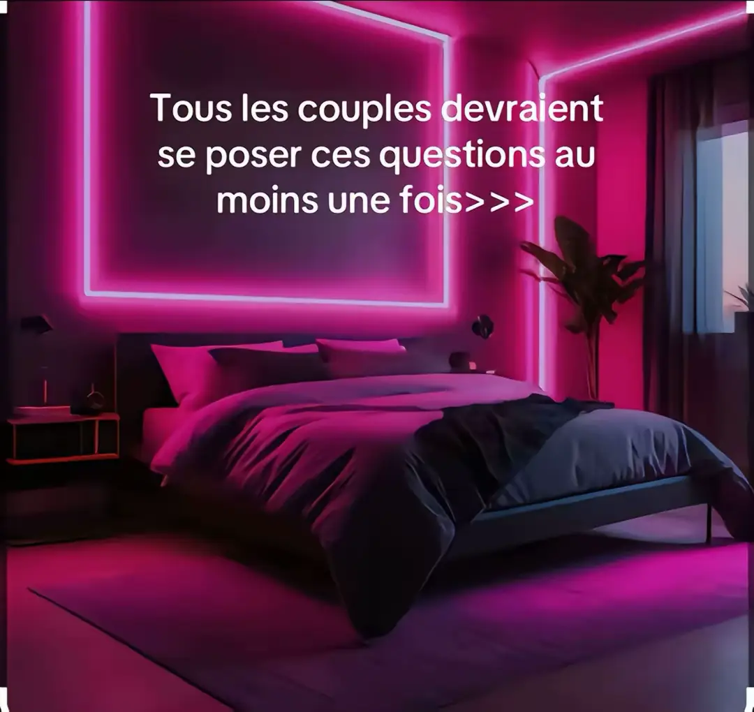 A bedroom interior with a modern, neon-lit design. The room is bathed in a magenta glow, created by large, illuminated rectangular frame on the wall and other light sources. The bed is the focal point, covered with a dark purple duvet and pillows. A side table with a lamp is visible, along with a large plant in the corner and a window.