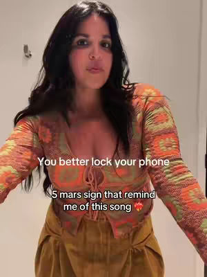 A woman with long black hair stands in a room, wearing an orange patterned top and a mustard-colored bottom. The top has a plunging neckline, and text overlayed on the image.