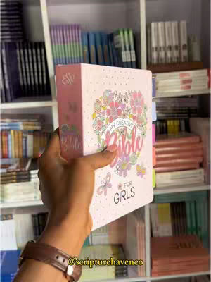 A person's hand holds a pink Bible with a floral design and the title 'My Creative Bible for Girls' in front of a bookshelf. The background shows various books, suggesting a bookstore or library setting.