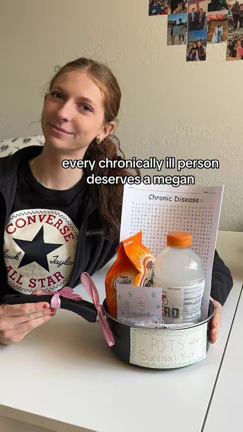 A young woman smiles at the camera, holding a black frying pan that contains a 'POTS Survival Kit.' Behind her is a plain wall with photos and a framed picture. Text overlays read, 'every chronically ill person deserves a megan.'