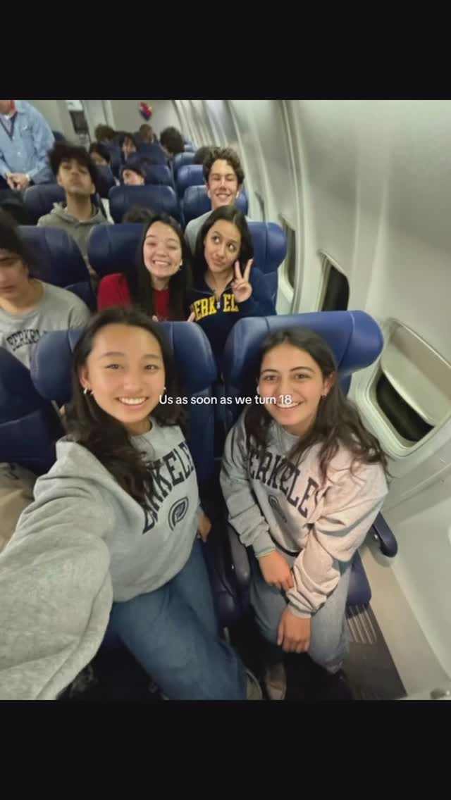 A group of young people posing inside an airplane. Two girls are in the foreground, one on the left taking a selfie with a smiling face, and another one on the right. The background is filled with a group of people in the seats, the image shows a sense of happiness and friendship. The text overlay says: Us as soon as we turn 18.