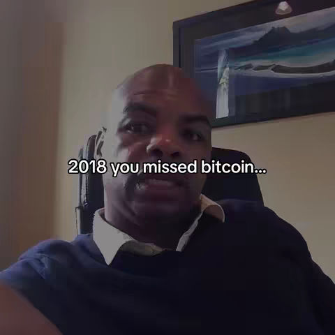 A man in a dark shirt sits in a chair, looking toward the camera. Text overlay says, '2018 you missed bitcoin...'. The background includes a framed piece of art and the wall is painted a flesh color.