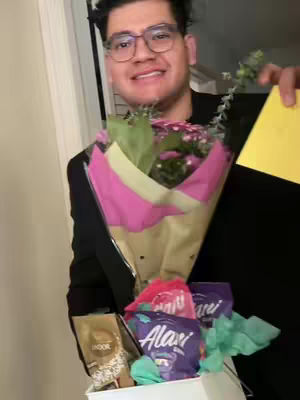 A man is holding a bouquet of flowers, an Easter basket filled with candies and other items, and a card. The man is wearing glasses and a black outfit, and he is smiling with a bouquet of pink and purple flowers. The basket contains candies with the Alani Nu brand. The background is a blurry interior setting.