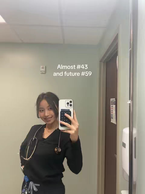 A selfie taken in a medical setting reflects in a mirror, which shows a woman in black scrubs holding her phone up, looking towards the camera, with a stethoscope around her neck. The background is simple, with a door and wall details.