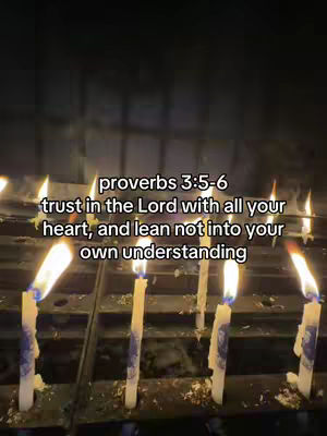 A low-angle shot of several lit candles on what appears to be a dark, possibly ornate surface. The background is out of focus but suggests a dimly lit or enclosed space. Text overlays are present, providing a religious quote.