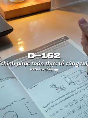 The image showcases an open textbook with handwritten mathematical equations and diagrams. On the top left corner of the book is a partially visible black laptop and a wooden surface. The overall composition suggests an educational theme, possibly a study or tutorial.