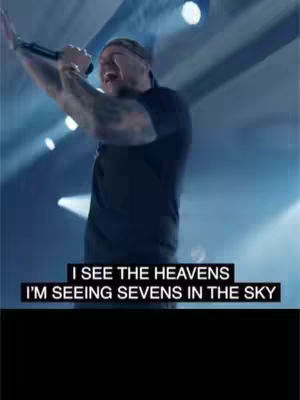 The image features a man on stage, holding a microphone and singing. He is wearing a black shirt. Behind him, the background is illuminated with blue and white lights, and there are lights on the stage.